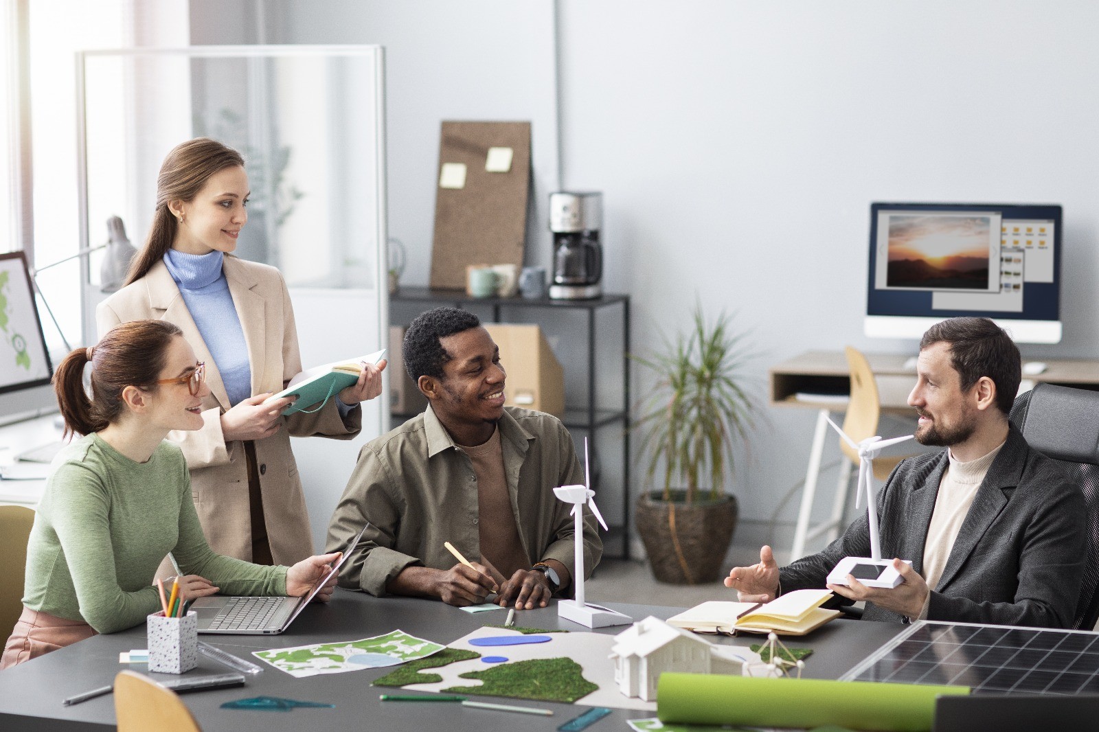 All'interno dell'immagine sono presenti quattro persone, due uomini e due donne. Tre sono seduti al tavolo a discutere di un progetto lavorativo, la quarta è in piedi che prende appunti.