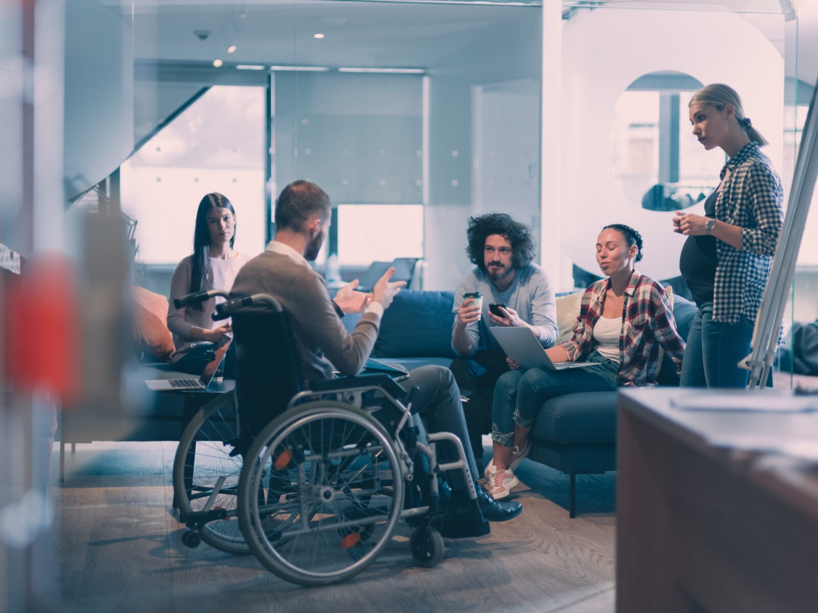 Da dove nasce questo vademecum: All'interno dell'immagine ci sono cinque persone che parlano in uno spazio comune. Tre sono sedute sopra un divano con il computer e il telefono. Una donna incinta è in piedi che ascolta attentamente un uomo in sedia a rotelle. Lo spazio comune si trova all'interno di un ufficio moderno